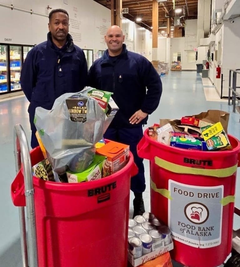 Two men standing in the Food Bank warehouse next to 2 large bins of donations on top of a dolly.