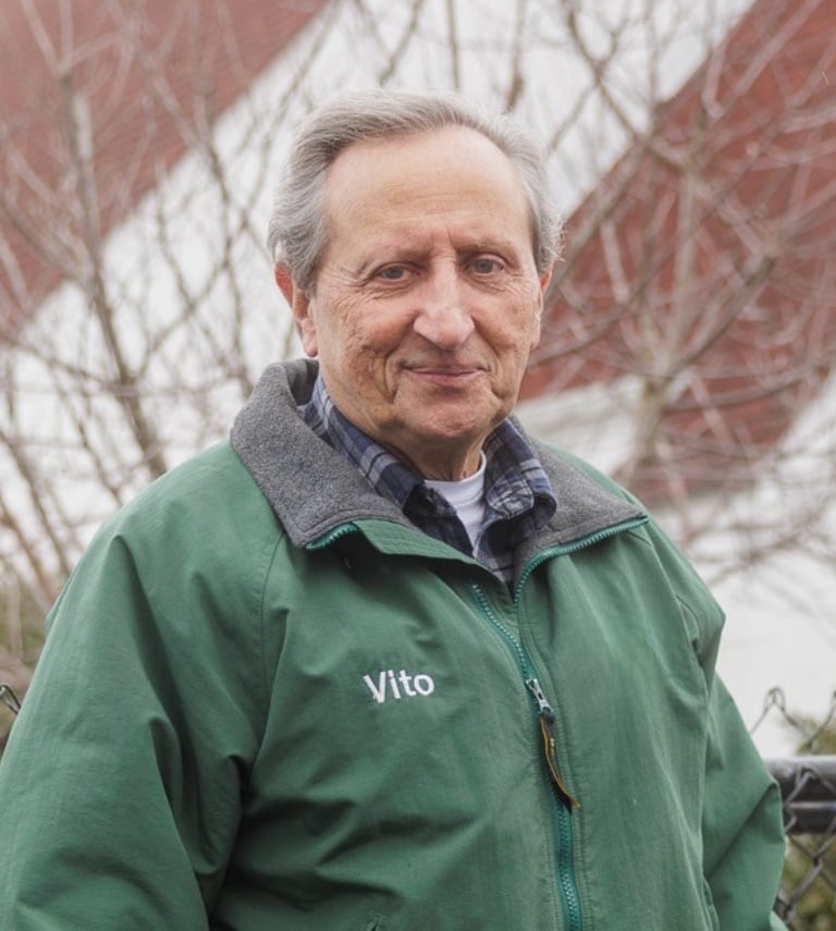 Older man standing outside and looking at the camera. He's wearing a winter coat with the word Vito on it.