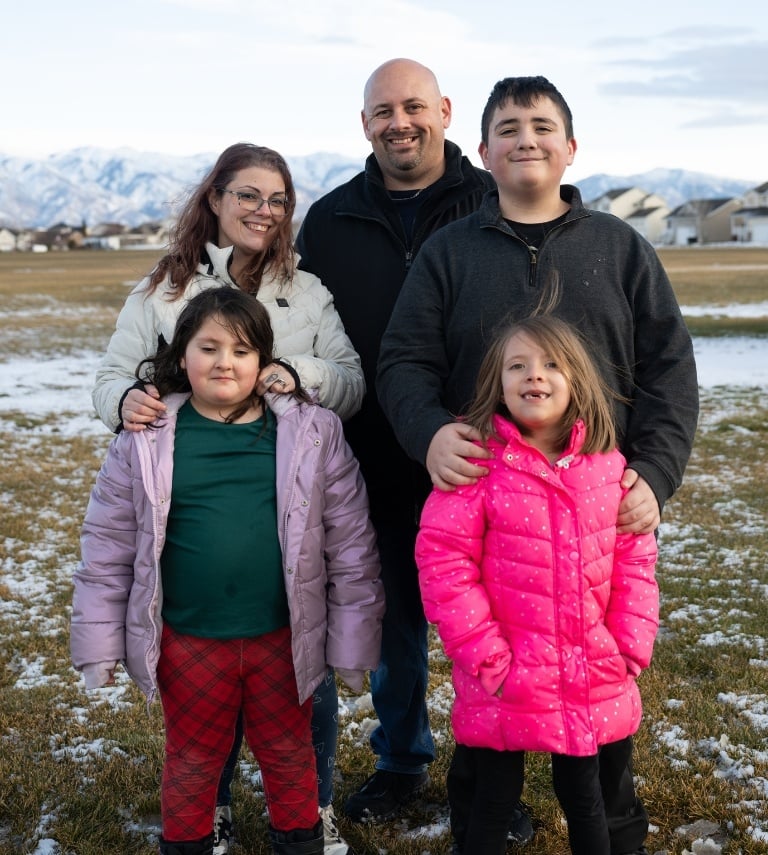 Family of 5 outside looking at the camera. There are two young girls, one teenage boy, a mother and a father.