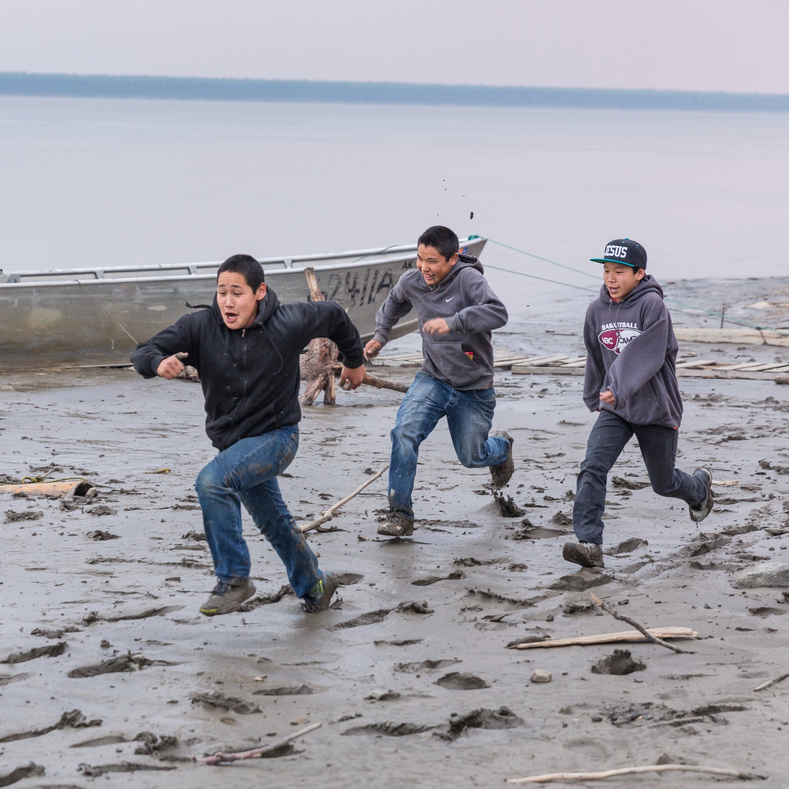 Three boys running on a beach in rural Alaska, alongside a boat