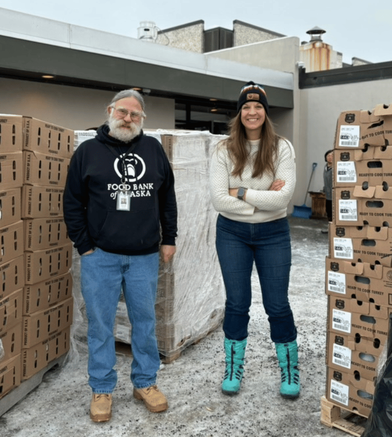 An older Food Bank employee and a younger food bank employee standing next to each other outside with pallets surrounding them