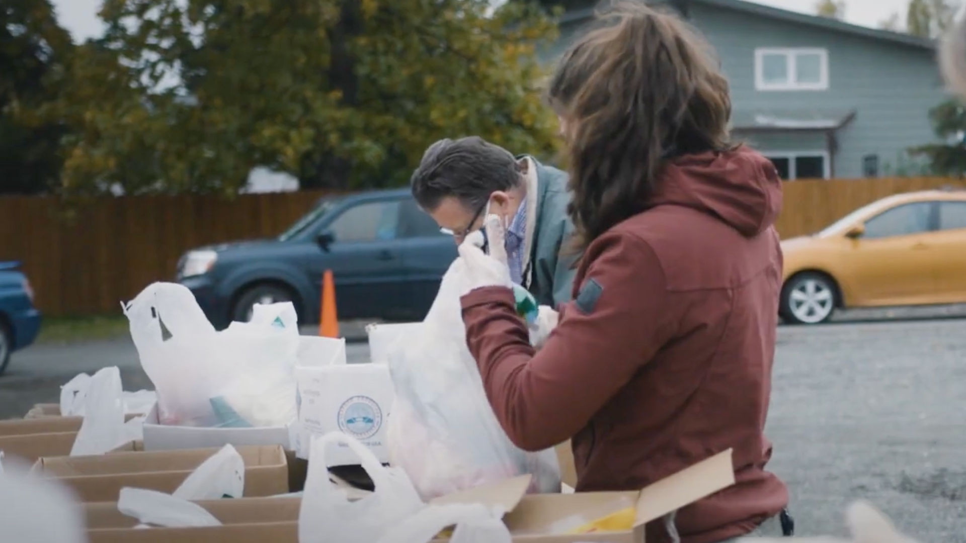 Man and woman facing away from camera, filling white plastic bags outside with produce from a table.